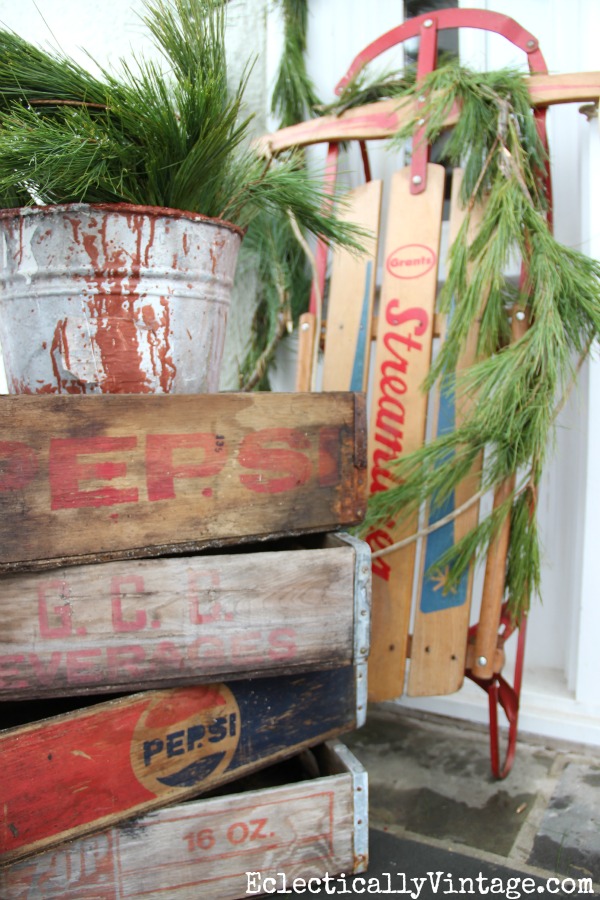 Love this stack of old wood soda crates used as a plant stand on this fun winter porch kellyelko.com #winterdecor #vintagechristmas #winterporch #christmasporch #vintagedecor #sodacrates #sled #kellyelko