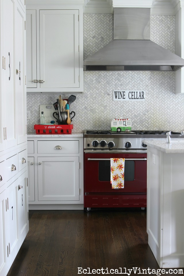 Love this white kitchen and carrara marble herringbone tile - the red stove really pops! kellyelko.com #spring #springdecor #springdecorating #springcrafts #hometour #housetour #eclectichome #farmhousedecor #fixerupperstyle #jadeite #farmhousestyle #vintagedecor #vintagehome #kellyelko #redstove #whitekitchen #backsplash #marbletile #herringbonetile #vintagedecor #vintagekitchen #kitchenreno