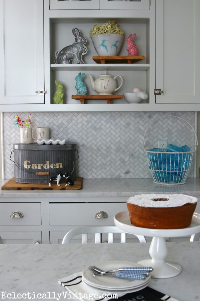 White spring kitchen - lots of fun display ideas and I love the galvanized garden pail kellyelko.com #spring #springdecor #springdecorating #springcrafts #hometour #housetour #eclectichome #farmhousedecor #fixerupperstyle #jadeite #farmhousestyle #vintagedecor #vintagehome #kellyelko #easterdecor #bunnies #whitekitchen #marblebacksplash #carraramarble