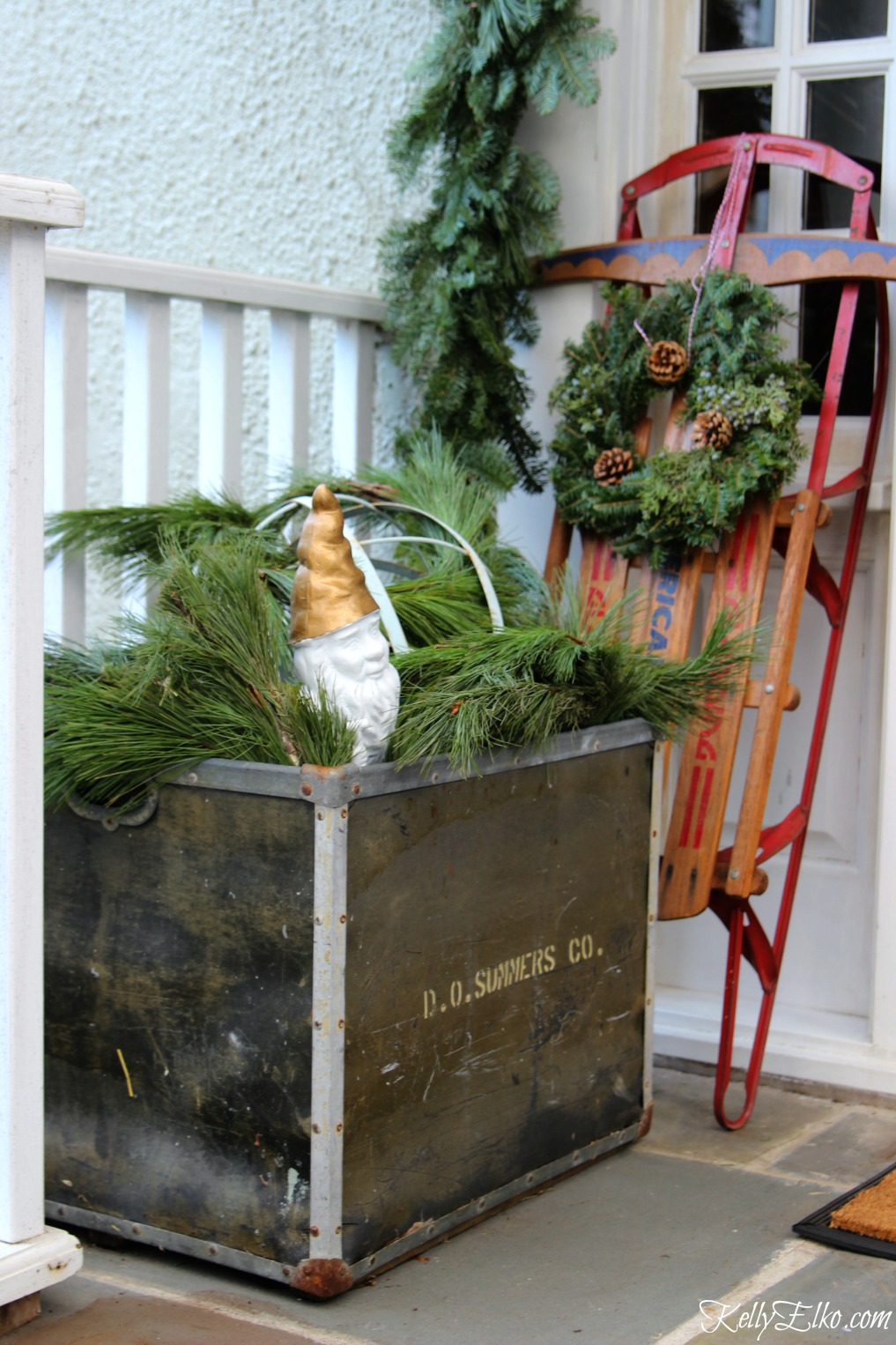 Vintage Christmas porch decorating ideas - love using a big metal container to hold sprigs of fresh greenery kellyelko.com #christmasporch #christmasdecor #outdoorchristmasdecor #vintagechristmas #sled #kellyelko #christmaswreath 