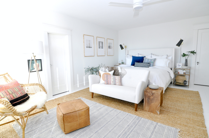 White master bedroom with texture from jute rug, leather pouf, rattan chair and wood accents