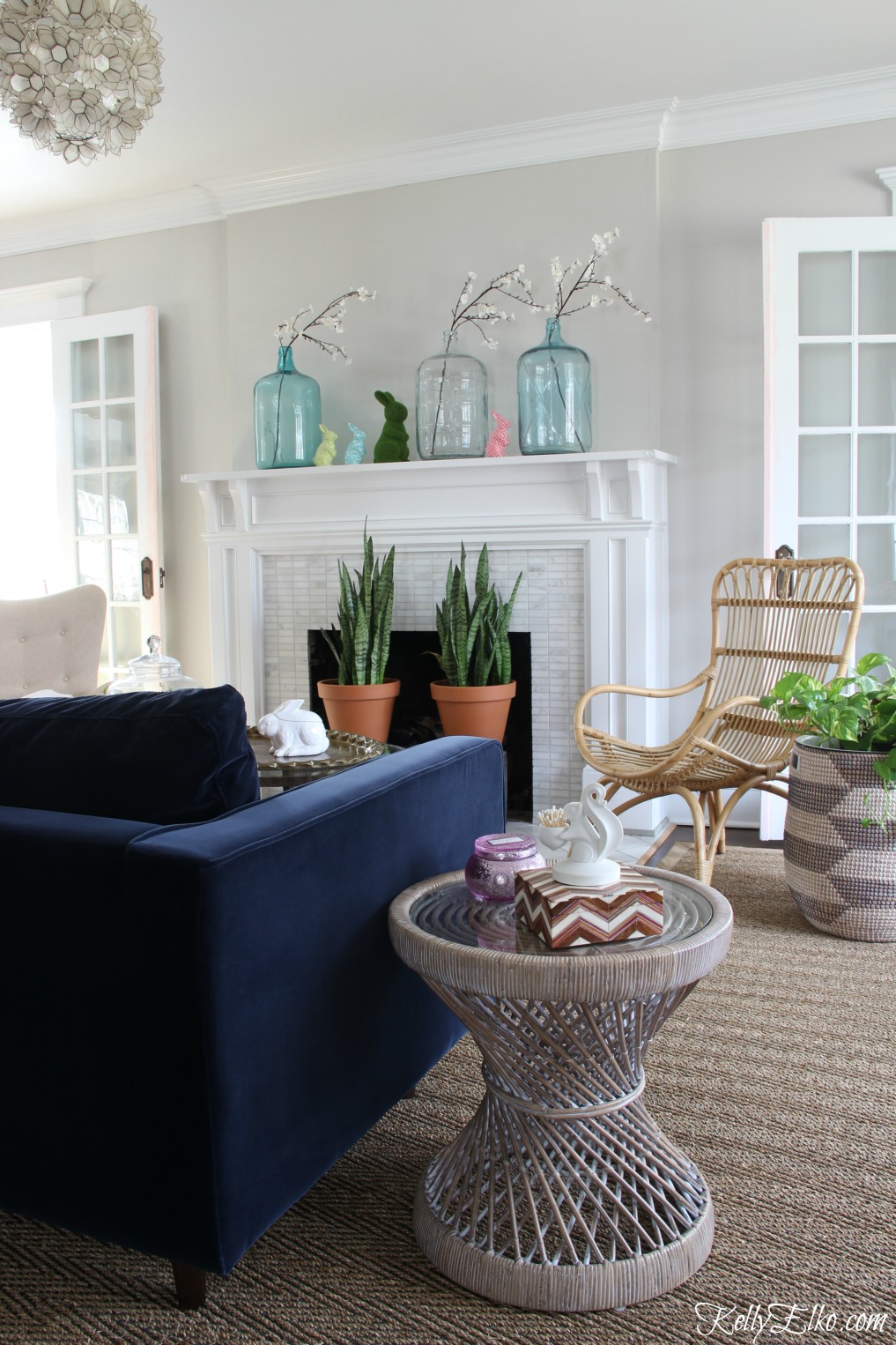 25 Spring Decorating Ideas - love this living room mantel with green glass jugs and bunnies. Rattan furniture, baskets and plants add a boho feel. kellyelko.com #spring #springdecor #homedecor #mantel #springmantel #vintagedecor