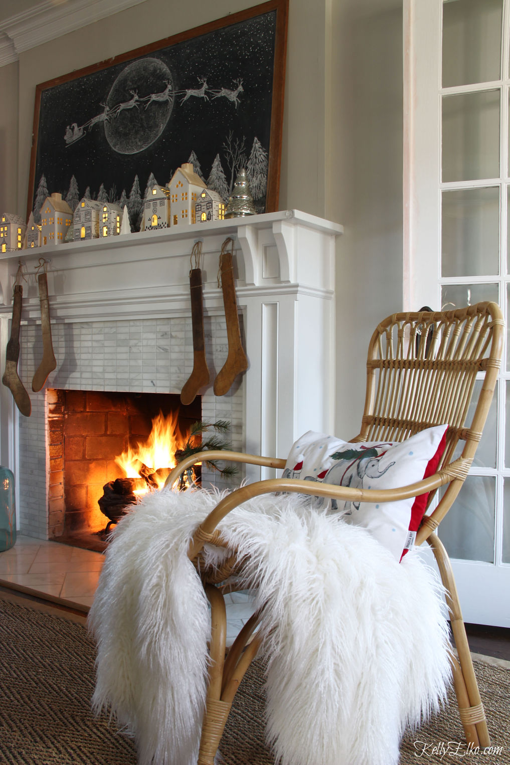 Love this chalkboard Christmas mantel with little light up houses and rattan chair with cozy throw kellyelko.com #christmasdecor #christmasmantel #chalkboard #christmaschalkboard #chalkboardart #farmhousechristmas #boho #bohochristmas #rattan #vintagechristmas #christmaslivingroom