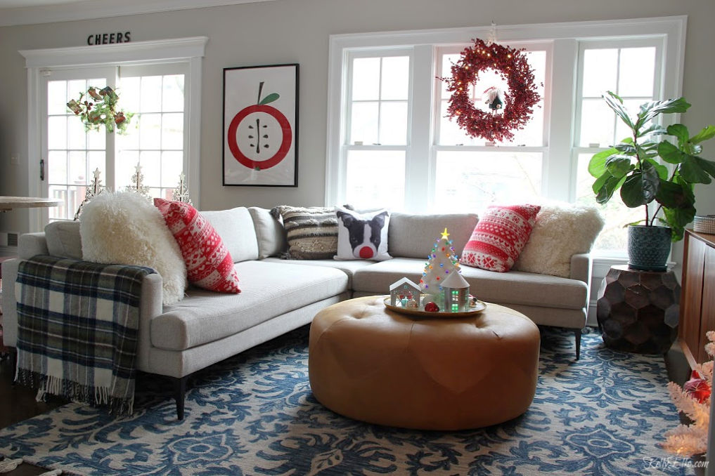 Cozy Christmas family room - love the Christmas dog pillow on the sectional sofa and huge berry wreath kellyelko.com #christmasdecor #christmaspillows #chritmaswreath #cozychristmas #leatherottoman #loloirug #modernart #fiddleleaffig