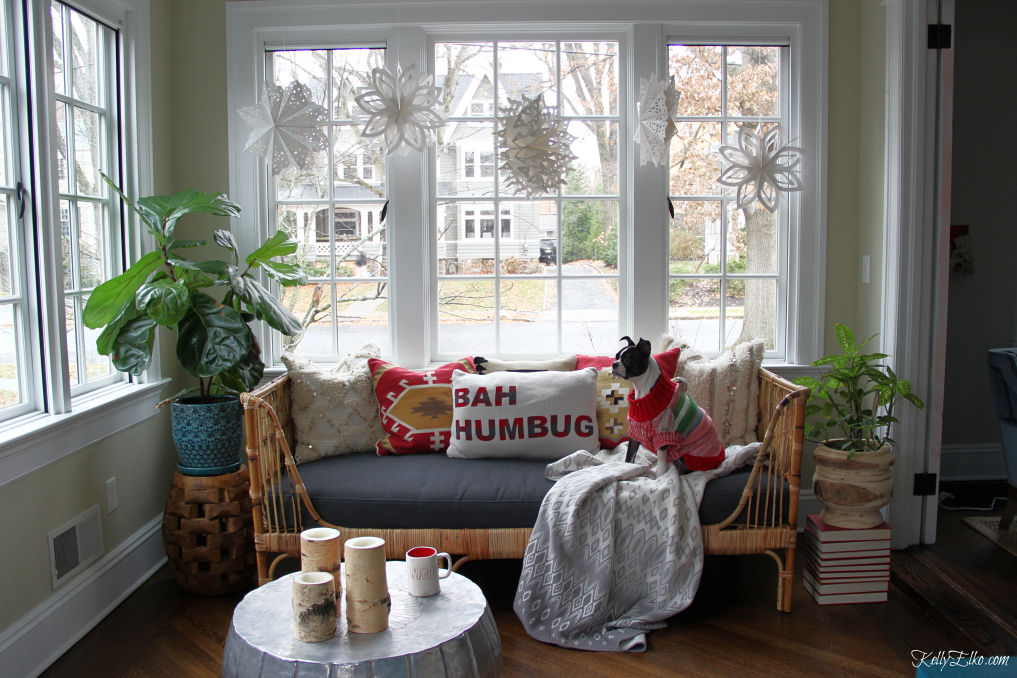 Love this sunroom decked out for Christmas with a boho rattan daybed covered in colorful pillows and cute paper snowflakes in the window kellyelko.com #bohodecor #bohodecorating #rattan #daybeds #sunroom #sunroomdecor #plantlady #houseplants #interiordecorate #interiordecor #christmas #christmasdecor #bostonterrier 