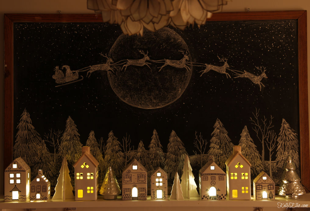 Beautiful Christmas mantel at night - love the way the little ceramic houses glow against the chalkboard art background kellyelko.com #christmas #christmslights #christmasdecor #christmasdecorating #christmasdiningroom #vintagechristmas #retrochristmas #christmasart #chalkboard #chalkboardart #christmasvillage #christmamantel