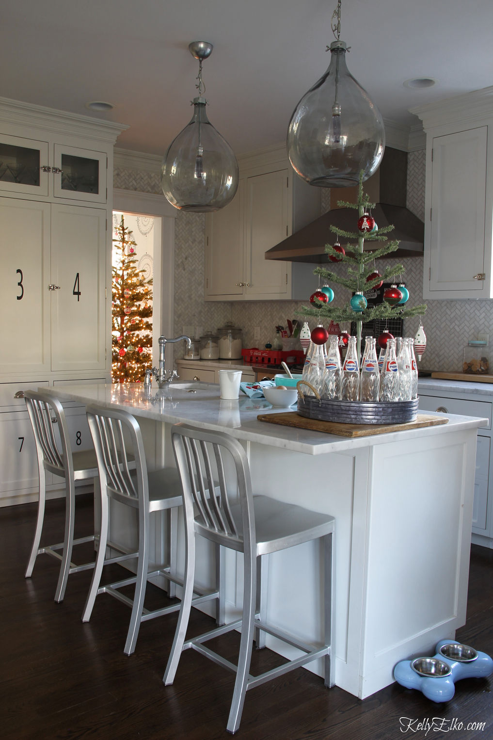 Beautiful white kitchen decorated for Christmas - love the demijohn lights kellyelko.com #christmaskitchen #farmhousekitchen #christmastree #lighting #kitchenisland #christmasdecor #christmasdecorating 