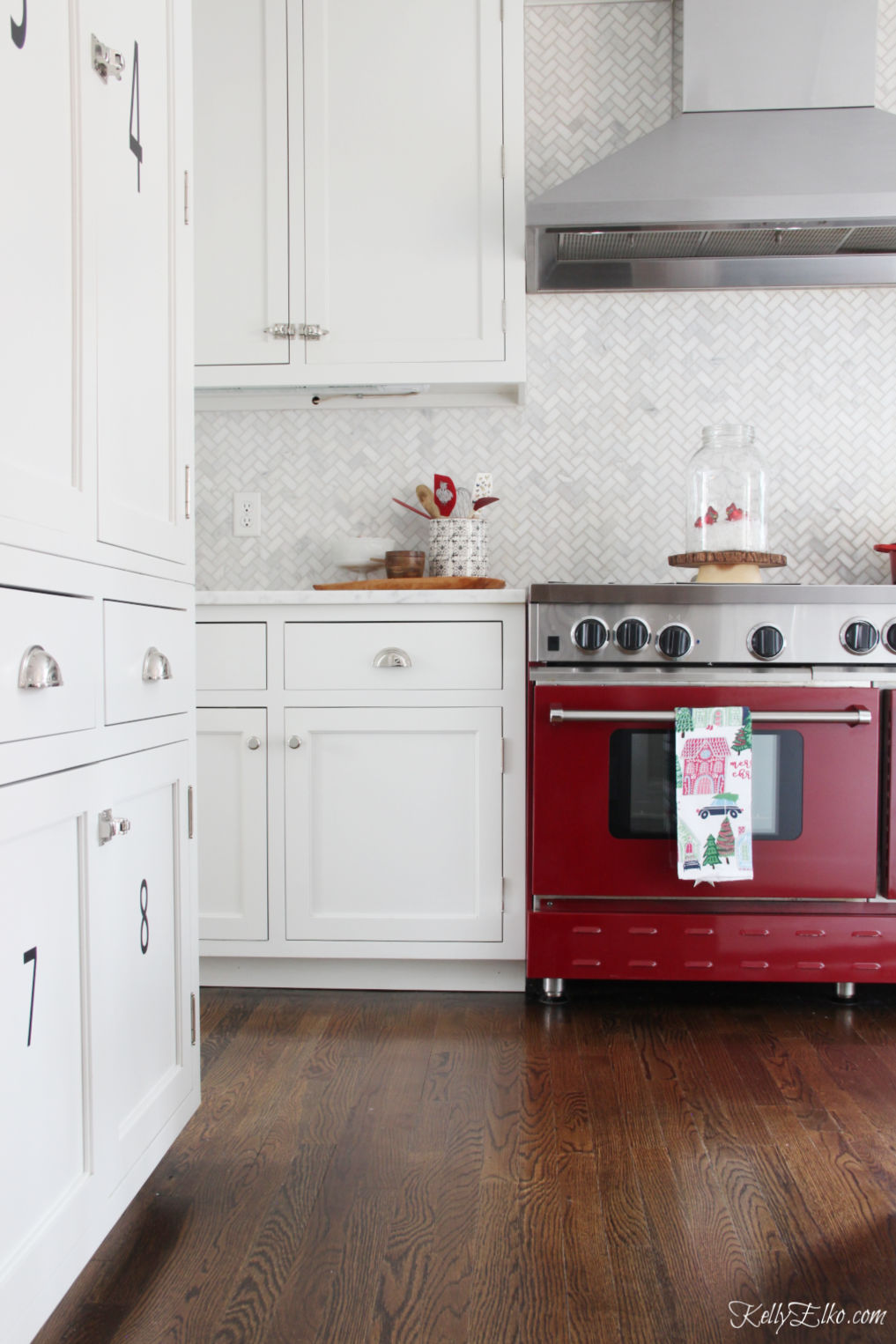 Beautiful white kitchen with red stove, carrara herringbone tile and white cabinets kellyelko.com #christmas #christmasdecor #christmasdecorating #christmashome #christmastour #diychristmas #christmasideas #christmasmantel #christmastree #christmasornaments #vintagechristmas #farmhousechristmas #colorfulchristmas #creativechristmas #kellyelko #redstove #stove #backsplash #whitekitchen 