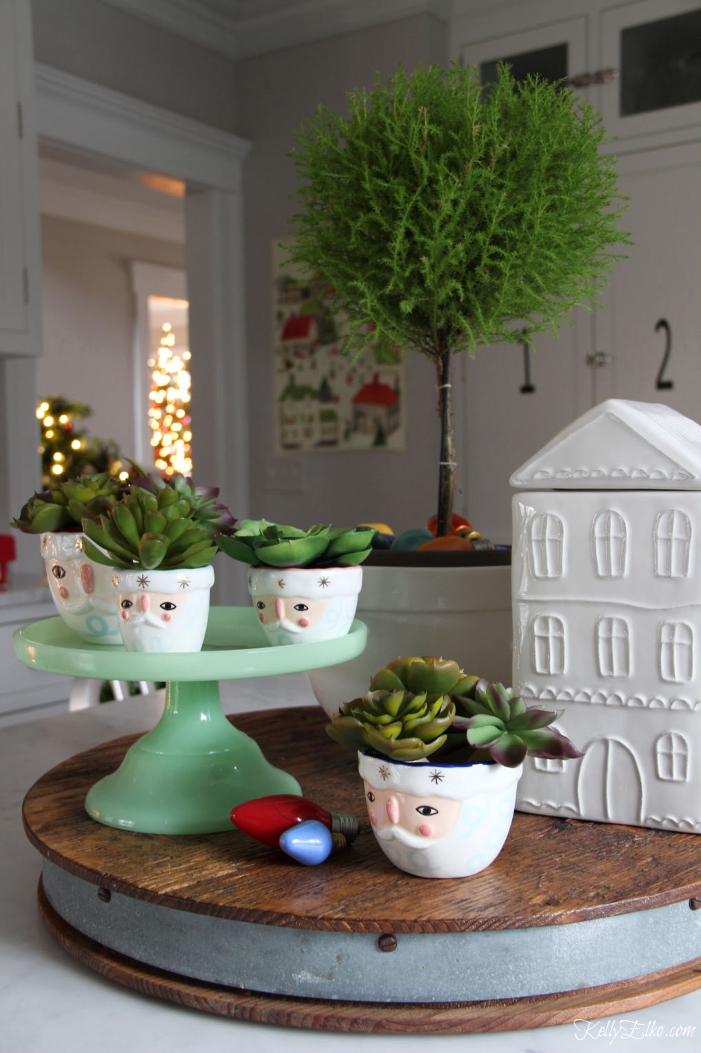 Cute kitchen Christmas vignette of Santa measuring cups, house cookie jar and a beautiful topiary kellyelko.com #christmas #christmasdecor #christmasdecorating #christmashome #christmastour #diychristmas #christmasideas #christmasmantel #christmastree #christmasornaments #vintagechristmas #farmhousechristmas #colorfulchristmas #creativechristmas #kellyelko #topiary #santas #santadecor #jadeite 