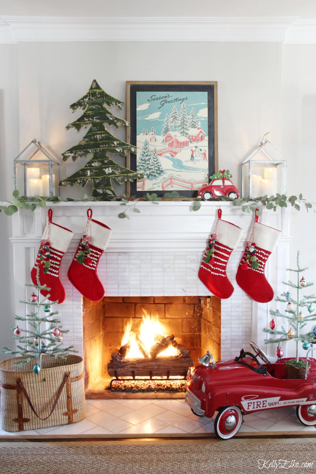 Love this vintage Christmas mantel with retro art and cute little red fire truck with feather trees kellyelko.com #christmas #christmasdecor #christmasmantel #retrochristmas #vintagechristmas #farmhousechristmas #fireplace #cozychristmas #feathertrees #christmastree #christmasstockings #eclecticchristmas