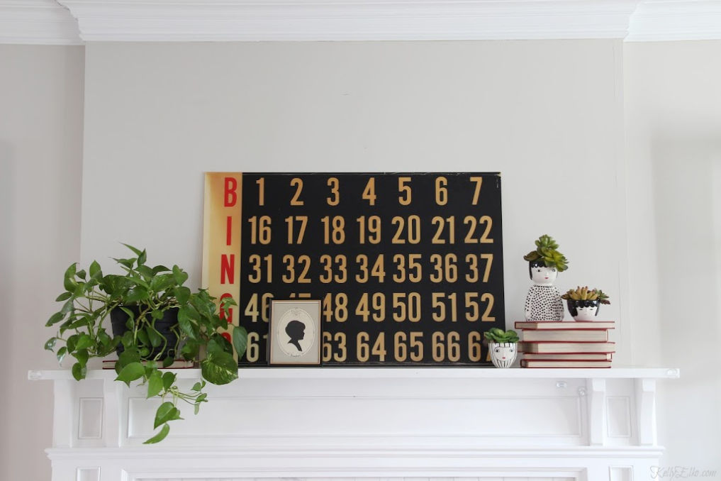 Love this eclectic vintage modern mantel with an old Bingo scoreboard, plants and face planters kellyelko.com #vintagemodern #vintagedecor #farmhousedecor #fixerupperstyle #plantlady #pothos #houseplants #succulents #thrifteddecor #thrifted #thrifty #kellyelko 