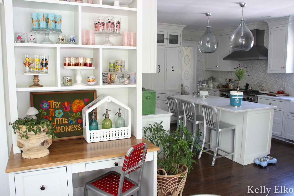 Love this colorful vintage drinking glass display in this white kitchen with farmhouse plate rack and bubble chandelier kellyelko.com #vintageglass #vintageglasses #drinkingglasses #vintagedrinkingglass #collectibles #collections #collect #collector #kitchendecor #vintagedecor #colorfuldecor #thrifted #vintagemodern #shelfie #kellyelko #kitchen #platerack #mugrack #farmhousedecor #farmhousekitchen