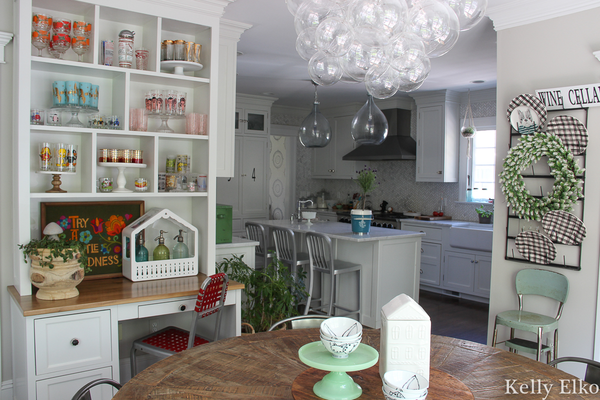 Love this colorful vintage drinking glass display in this white kitchen with farmhouse plate rack and bubble chandelier kellyelko.com #vintageglass #vintageglasses #drinkingglasses #vintagedrinkingglass #collectibles #collections #collect #collector #kitchendecor #vintagedecor #colorfuldecor #thrifted #vintagemodern #shelfie #kellyelko #kitchen #platerack #mugrack #farmhousedecor #farmhousekitchen