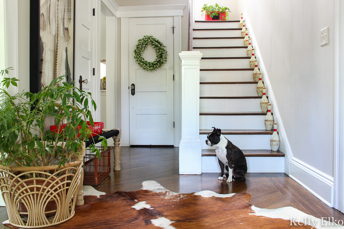 Beautiful foyer with vintage bowling pins on the stairs and fun pops of red kellyelko.com #foyer #foyerdecor #entry #entrydecor #farmhouse #farmhousedecor #vintage #vintagedecor #collections #collect #cowhide #plants #plantlady #houseplants #bohodecor #boho #eclectic #eclecticdecor 