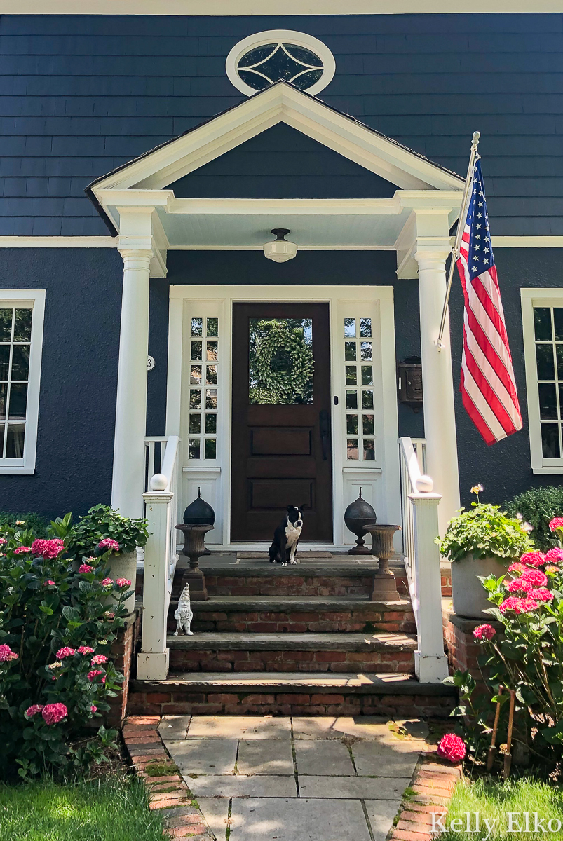 Love this bold blue house and charming small front porch kellyelko.com #blue #bluepaint #bluehouse #halenavy #housepaint #housecolors #curbappeal #oldhouse #oldhome #oldhouses #fixerupper #frontporch #landscaping #gardening #gardens #dormers #farmhouse #farmhousedecor #flag #americanflag #hydrangeas #porchdecor