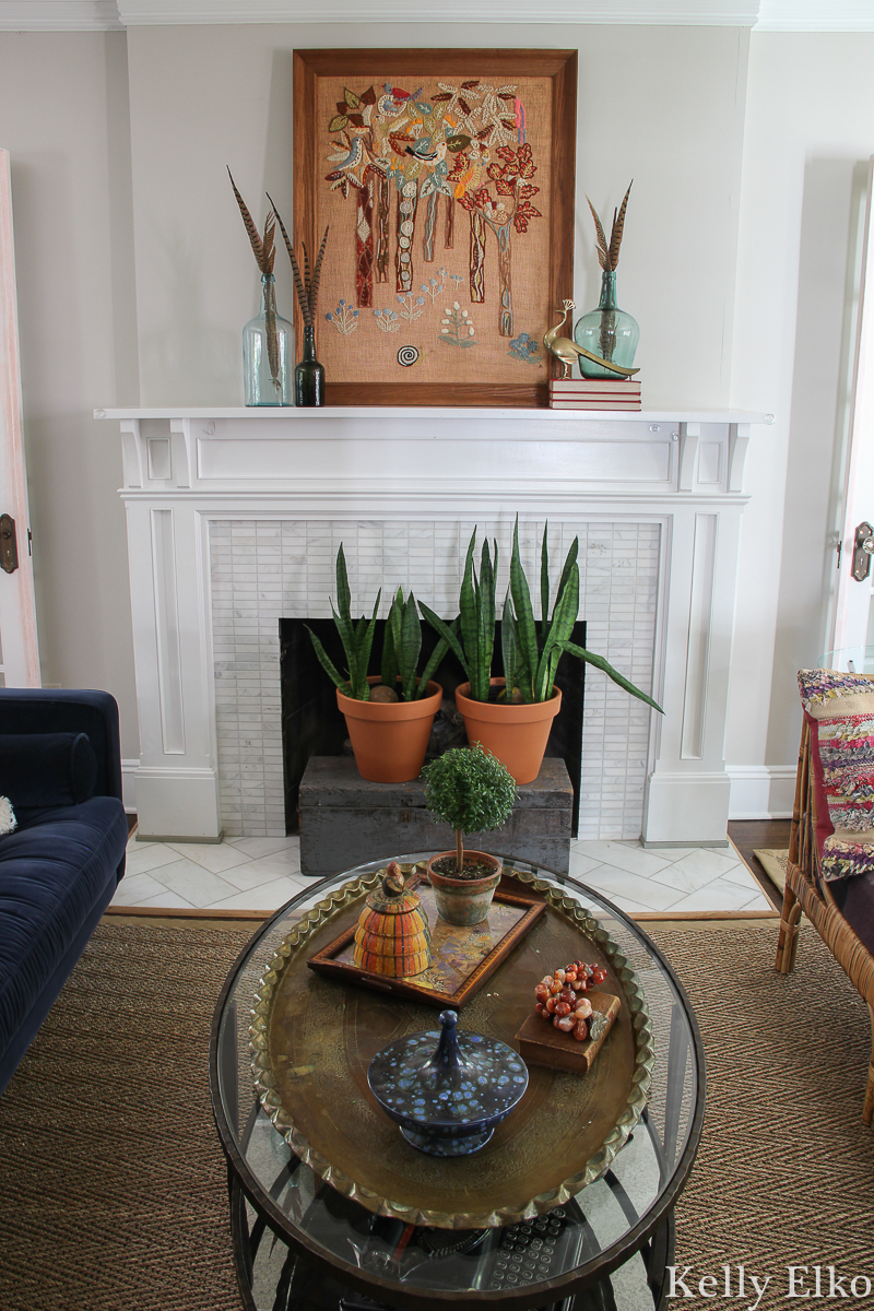 Love this eclectic living room with vintage crewel art and other unique pieces kellyelko.com #fall #falldecor #eclecticdecor #uniquedecor #livingroom #livingroomdecor #fallmantel #manteldecor #vintageart #crewel #houseplants #hearth 