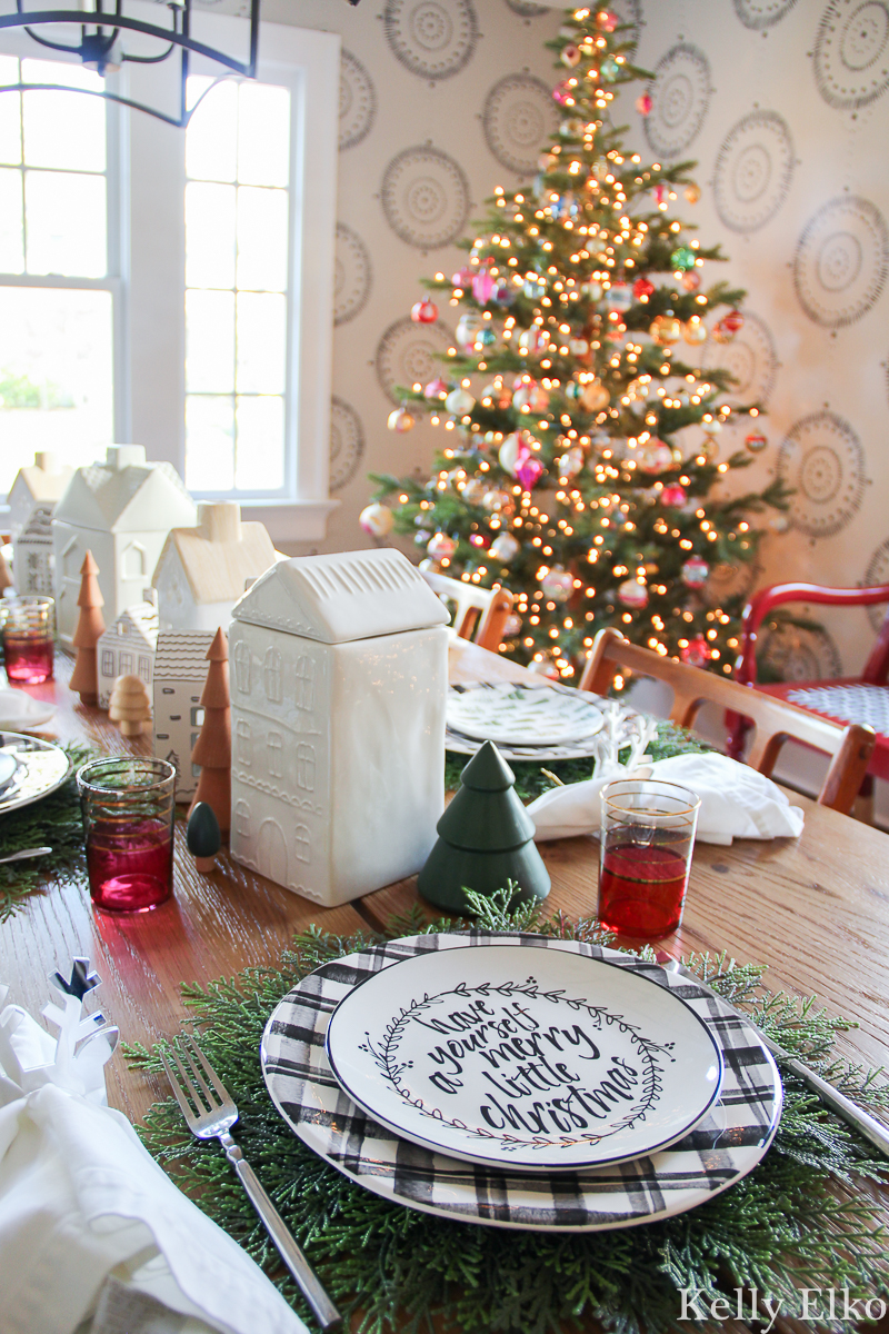 Love this festive Christmas table setting with plaid plates and cedar placemats and the cute little ceramic house centerpiece kellyelko.com #christmasdecor #christmastable #christmasplates #christmascenterpiece #christmasdiningroom #farmhousechristmas #christmastree #vintagechristmas 