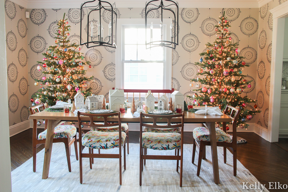 Love this beautiful Christmas dining room with a pair of realistic Christmas trees against a wall mural kellyelko.com #wallmural #christmasdiningroom #christmasdecor #vintagechristmas #farmhousechristmas #eclecticchristmas #sparsechristamstrees #realisticchristmastrees #danishmodern #MCMfurniture #colorfulchristmas 