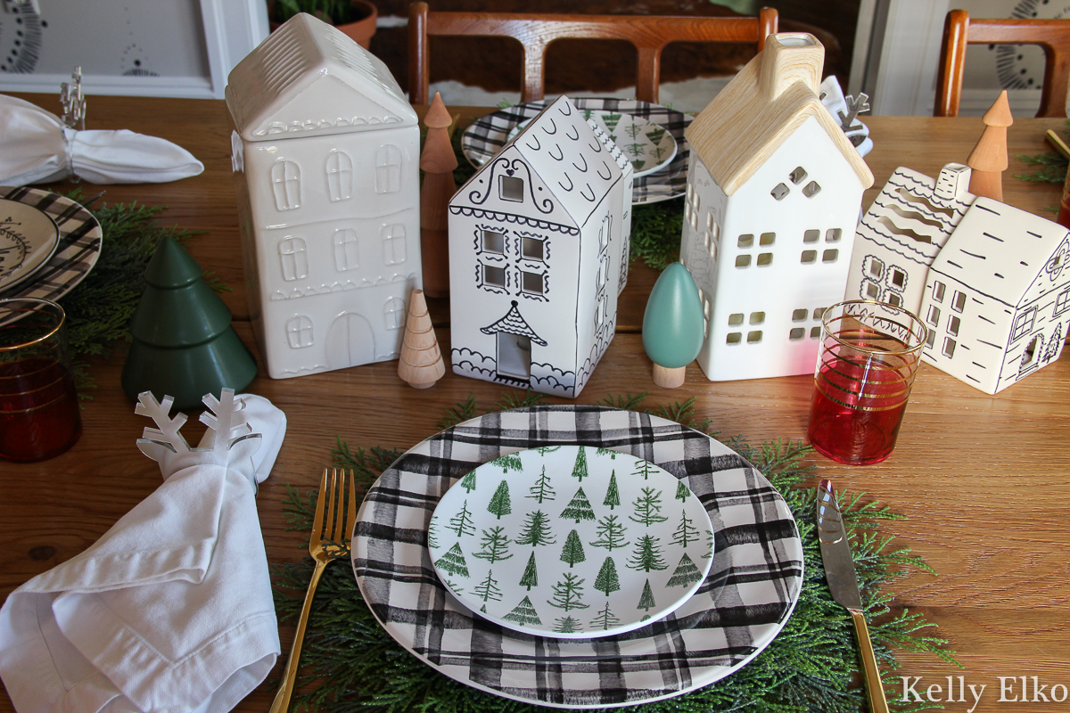 Love this little white ceramic Christmas houses centerpiece and the plaid plates with cute tree plates on top kellyelko.com #christmastable #christmascenterpiece #christmasplates #ceramichouses #whitechristmashouses #ceramicchristmashouses #diychristmasdecor #christmasplacesetting #farmhosuechristmas 