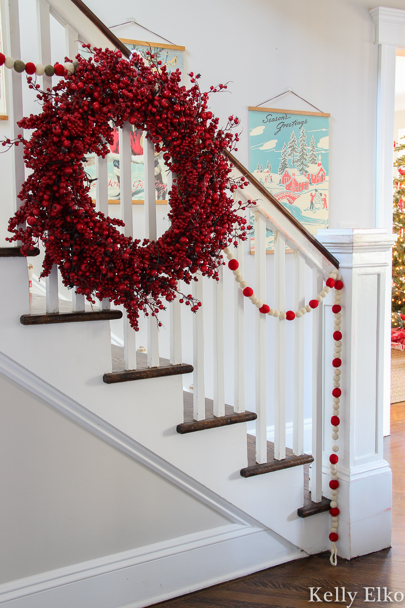 Love this retro Christmas art on the staircase wall in this beautiful home kellyelko.com #christmasart #vintageart #retroart #christmasfoyer #christmasentry #vintagechristmasart #retrochristmasart #vintagedecor #foyerdecor #entrydecor #berrywreath #christmasbannister #christmasgarland #farmhousechristmas