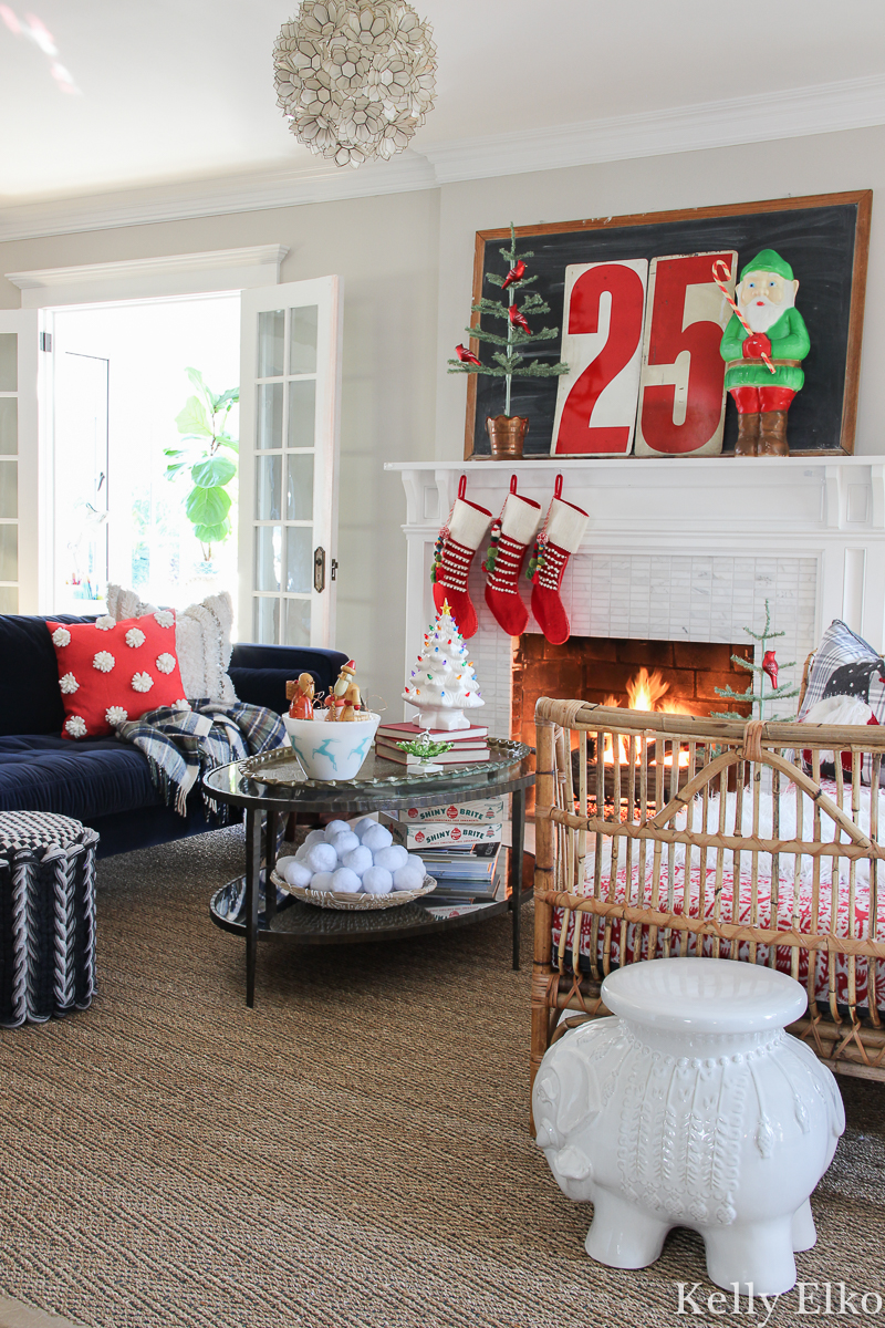 Love this colorful living room and the retro Christmas mantel with cute elf blow mold and pom pom stockings kellyelko.com #christmashouse #christmashometour #christmasmantel #retrochristmas #vintagechristmas #livingroomdecor #rattanfurniture #christmasstockings #blowmold #christmaslivingroom