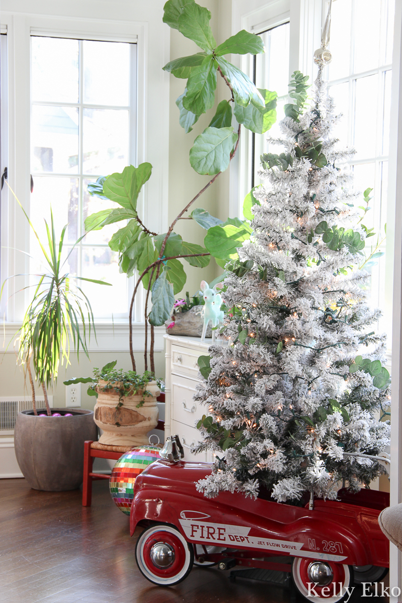 Love this retro fire truck pedal car turned Christmas tree skirt for a flocked tree kellyelko.com #christmasdecor #vintagechristmas #retrochristmas #farmhousechristmas #plants #sunroomdecor #fiddleleaffig #christmassunroom #flockedtree #pedalcar #vintagetoy