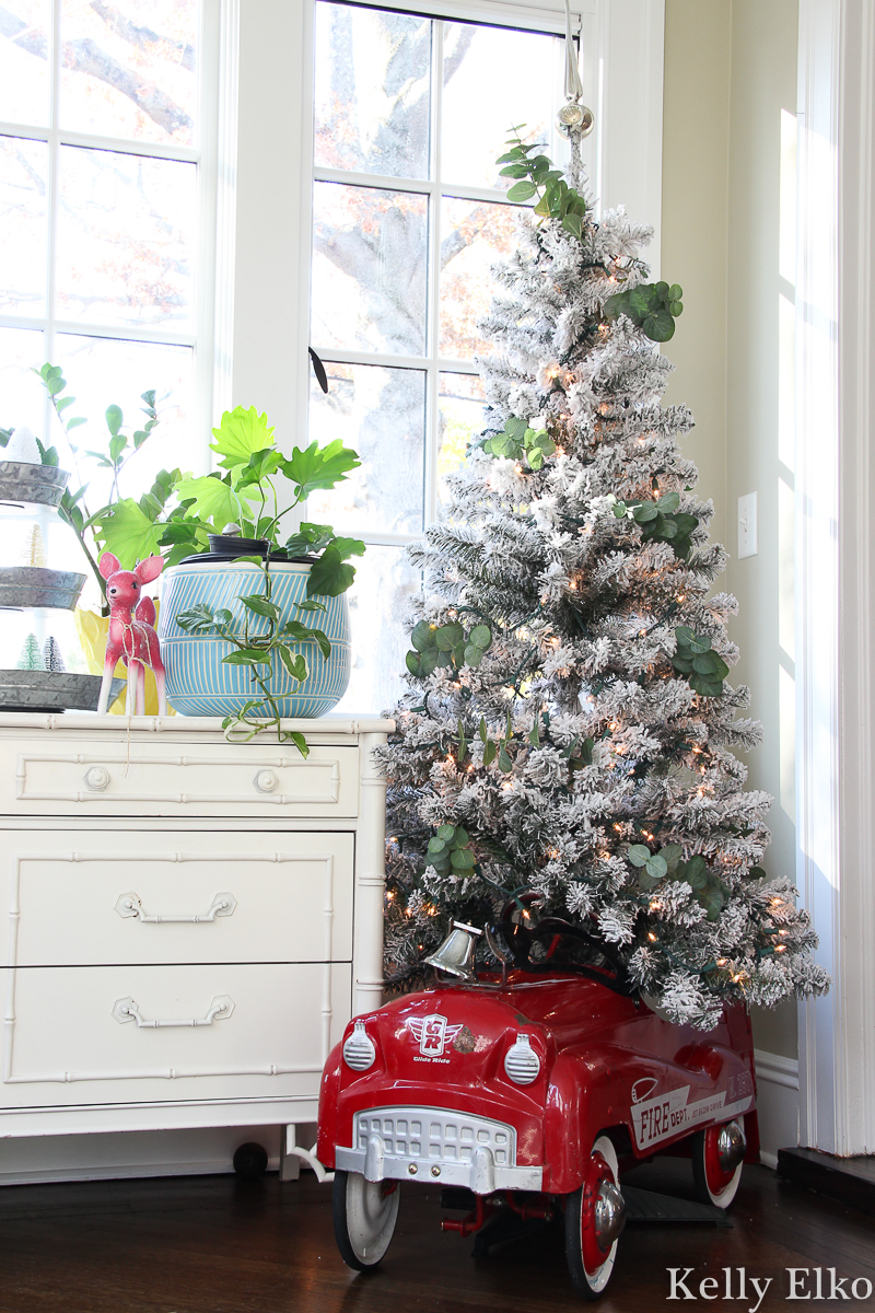 Love this retro fire truck pedal car turned Christmas tree skirt for a flocked tree kellyelko.com #christmasdecor #vintagechristmas #retrochristmas #farmhousechristmas #plants #sunroomdecor #fiddleleaffig #christmassunroom #flockedtree #pedalcar #vintagetoy