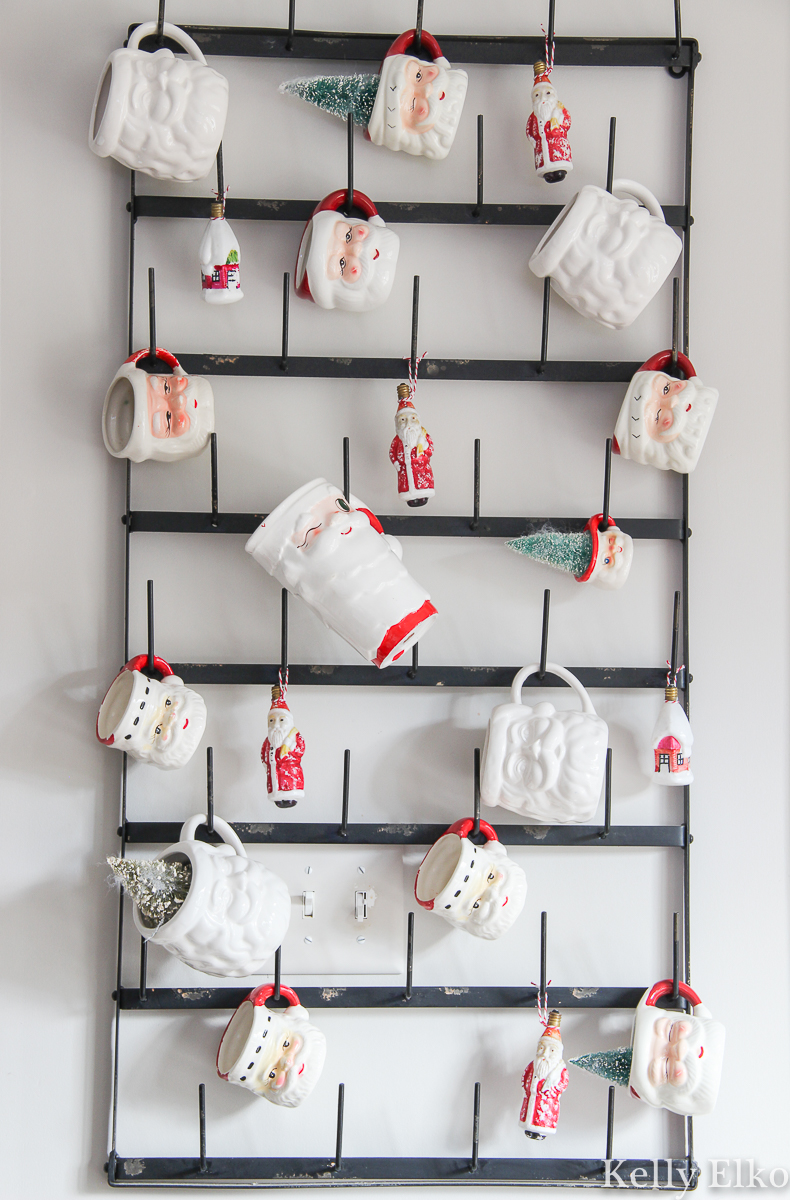 Love this display of Santa mugs hanging on a huge bottle drying rack kellyelko.com #santa #santamugs #vintagechristmas #vintagedecor #farmhousechristmas #christmasdecor #christmaskitchen #retrochristmas 