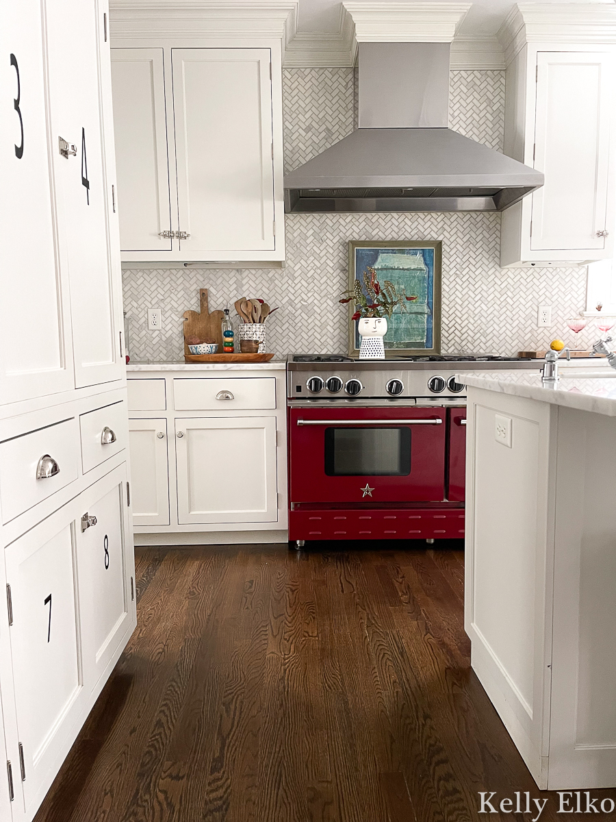 Love this farmhouse white kitchen with herringbone marble tile backsplash and stunning red BlueStar stove kellyelko.com