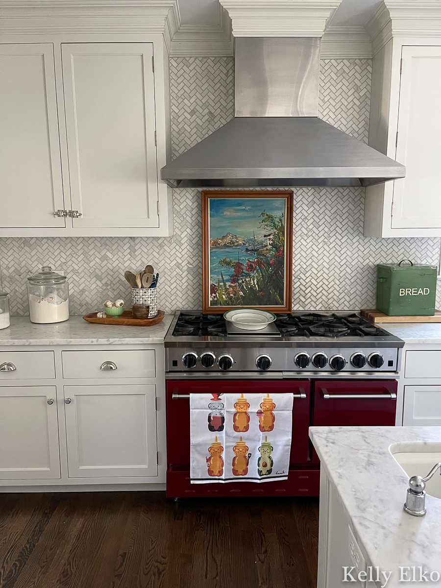 White kitchen with red stove and carrara marble herringbone backsplash and vintage art kellyelko.com